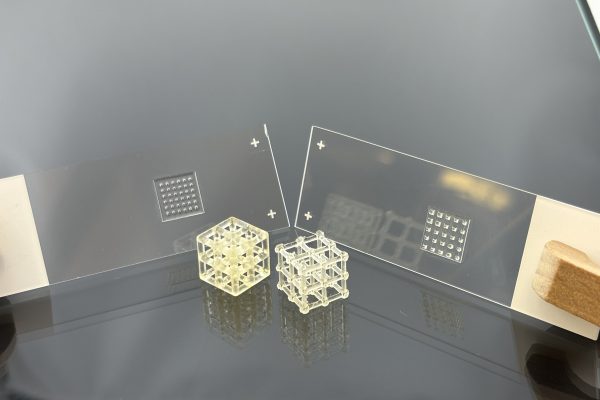 Transparente 3D-gedruckte Mikrostruktur-Gitterwürfel neben Lithografie-Masken auf Glas – Prototypen für Mikrooptik und additive Fertigung.