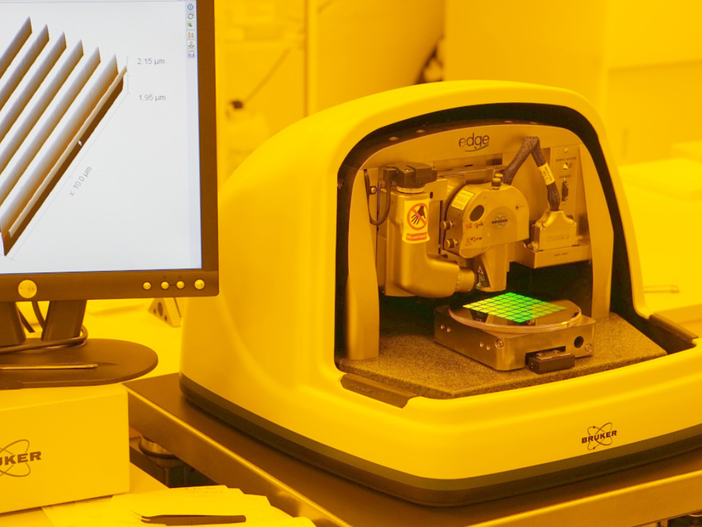 Laborgerät zur Oberflächenanalyse (Bruker Edge) in einem gelb beleuchteten Reinraum; im Inneren wird ein Wafer mit grün markiertem Messraster vermessen, während auf dem danebenstehenden Monitor eine vergrößerte 3D-Darstellung einer Mikrostruktur im Mikrometerbereich angezeigt wird.