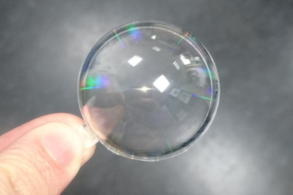 Hand hält eine transparente optische Linse mit nanostrukturierten Anti-Reflexionsoberflächen, die farbige Lichtreflexe zeigen – Beispiel für Nanoimprint-Lithographie in der Herstellung moderner optischer Komponenten.