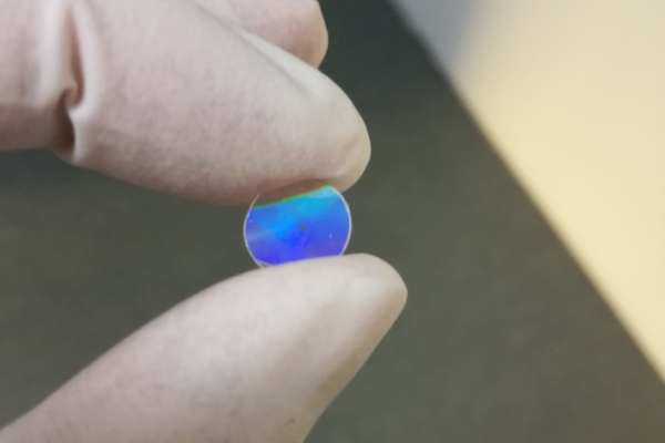 Kleines transparentes Bauteil mit schillernder nanostrukturierter Oberfläche, gehalten zwischen Fingern – Beispiel für Nanoimprint-Lithographie zur Herstellung funktionaler Nanostrukturen für Optik und Design.